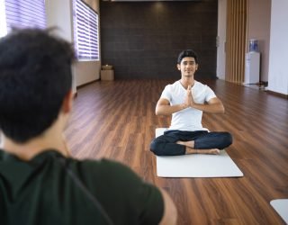 Yoga instructor meditating with student in gym. Man sitting in lotus pose and holding hands together with other guy sitting back to camera. Yoga class concept.